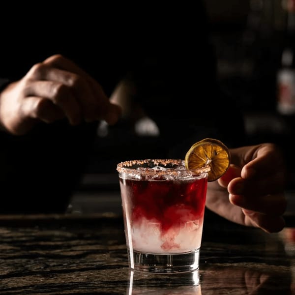 A bartender's hands prepare a red and white cocktail with a dried orange slice on a dark bar.