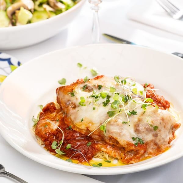 Chicken parmesan with melted mozzarella, red sauce, and microgreens, served with pasta.