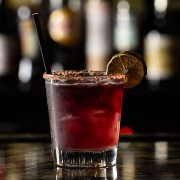 A red cocktail with a salted rim, lime slice, and black straw on a dark bar.