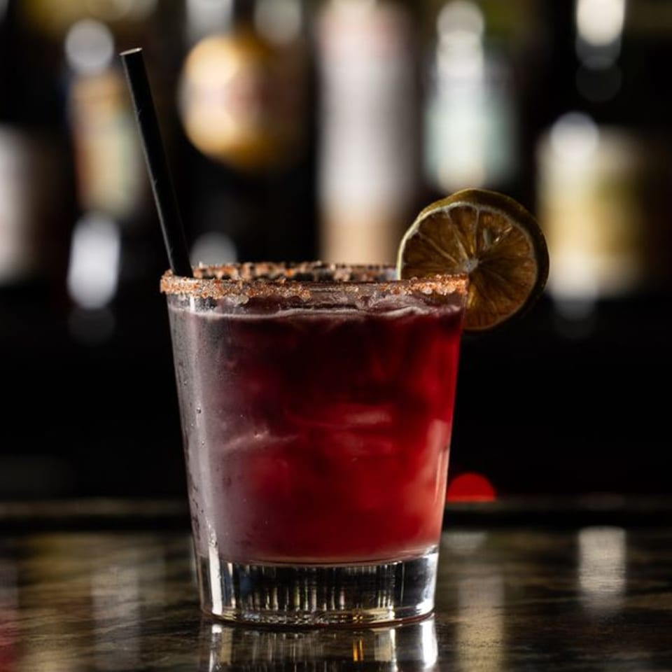 A red cocktail with a salted rim, lime slice, and black straw on a dark bar.