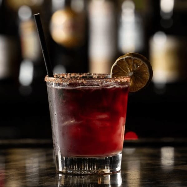 A vibrant red cocktail with a sugar-rimmed glass and a dried orange slice, on a dark bar.