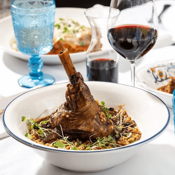 Braised lamb shank with risotto, garnished with microgreens, alongside red wine and a blue glass.