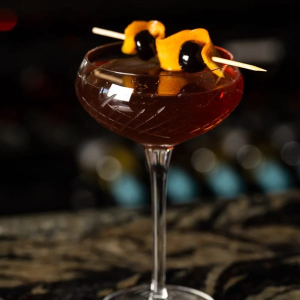 A dark cocktail in a coupe glass, garnished with orange peel and cherries on a skewer.