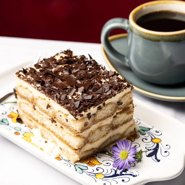 A slice of tiramisu with chocolate shavings and a purple flower on a decorative plate with coffee.