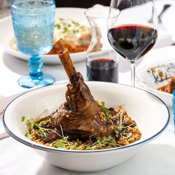 Braised lamb shank with rice and microgreens, alongside red wine and a blue glass.