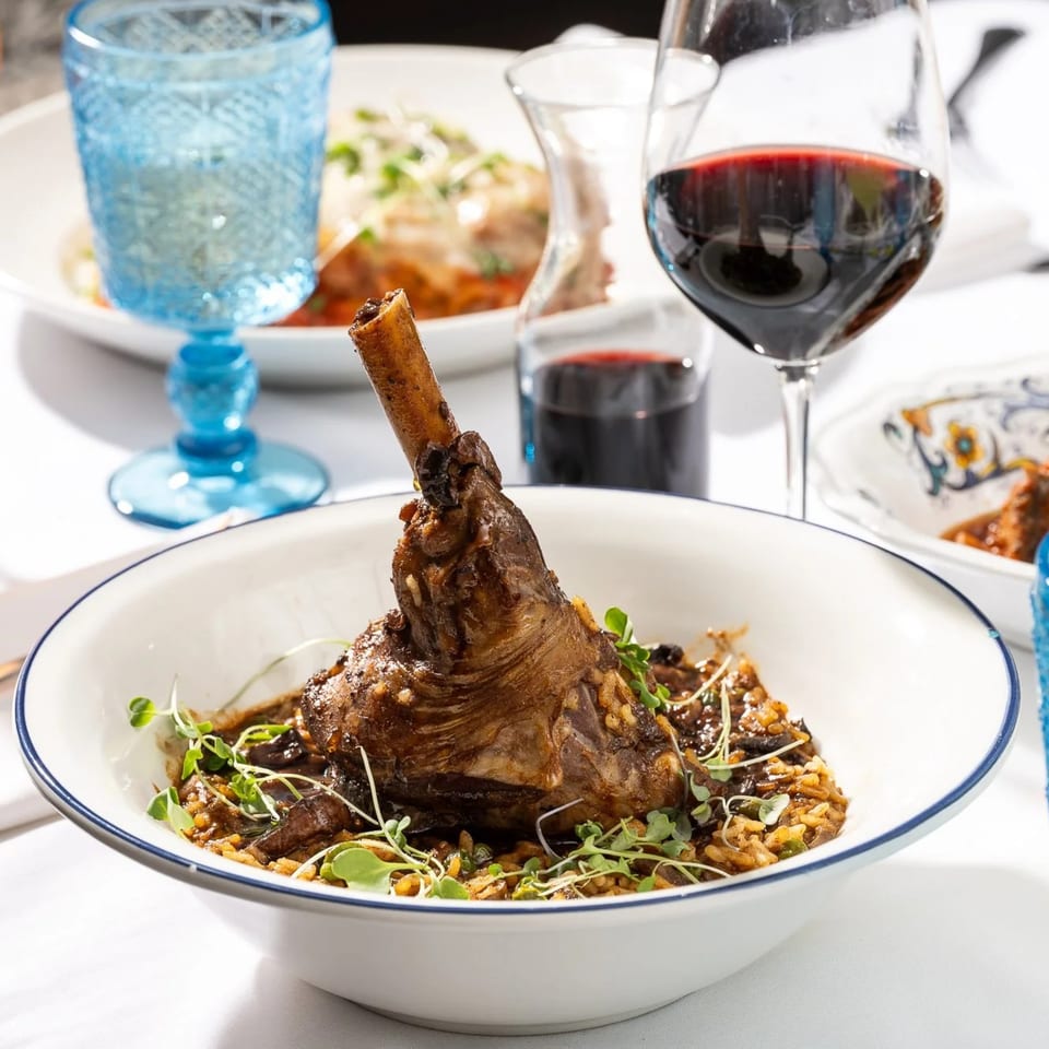 Braised lamb shank with rice and microgreens, alongside red wine and a blue glass.
