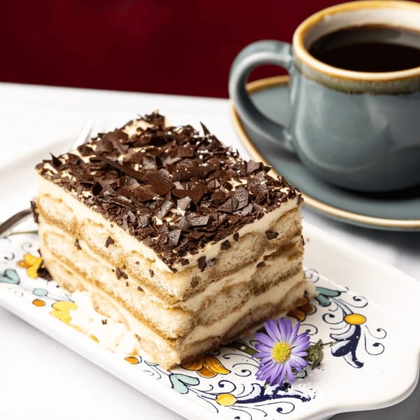A slice of tiramisu with chocolate shavings and a purple flower on a decorative plate next to a cup of coffee.